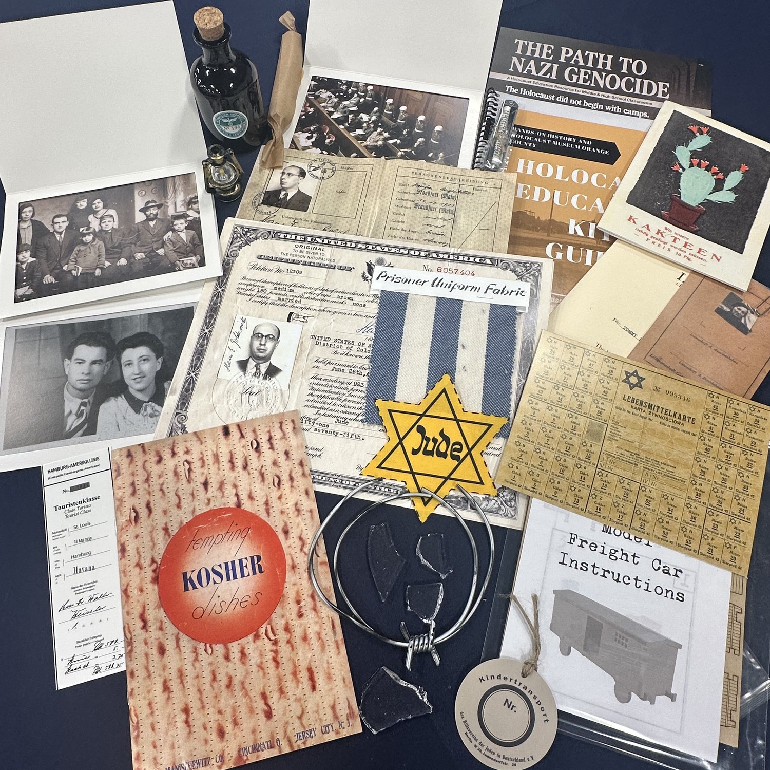 The contents of one Holocaust education kit laid out: family photographs, a German passport stamped with a red J, a United States naturalization certificate, a swatch of prisoner uniform fabric, a yellow star, a ghetto ration card, a Kindertransport tag, a shard of glass, a St. Louis boarding pass, a Kosher recipe pamphlet and the educator guide